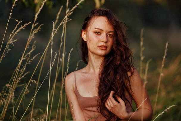 Beautiful woman face stand against wild grass, forest and bush in sunny rays field at sunset in summer meadow. Alone Cute young adult woman enjoy golden hou warm evening - Foto, afbeelding