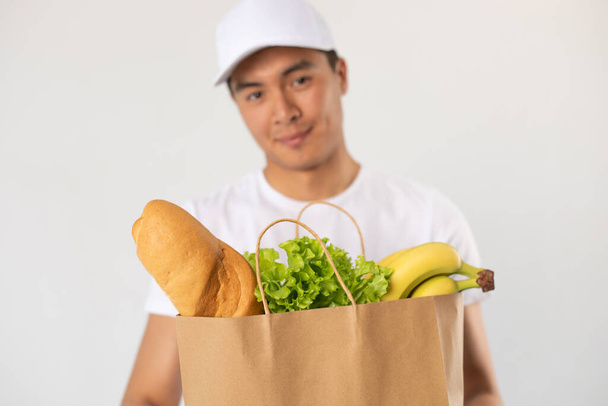 Asian delivery man in white cap holds craft bag with groceries on white background. - Фото, зображення