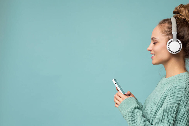 Side-ptofile closeup φωτογραφία της όμορφης νεαρής γυναίκας που φοράει κομψό casual ντύσιμο απομονωμένη πάνω από πολύχρωμο φόντο τοίχο φορώντας λευκά ασύρματα ακουστικά και ακούγοντας μουσική και χρησιμοποιώντας κινητό τηλέφωνο - Φωτογραφία, εικόνα