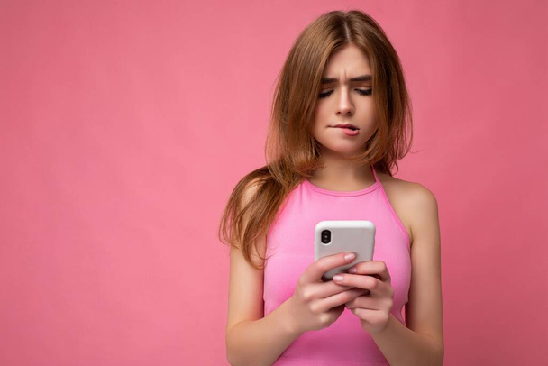 Detailní fotografie krásné přemýšlivé mladé blondýny na sobě růžové top balising izolované na růžovém pozadí s prázdným prostorem drží v ruce a pomocí mobilního telefonu surfování na internetu on-line - Fotografie, Obrázek