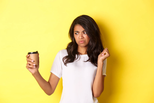 Портрет африканско-американской девушки, выглядящей разочарованной в чашке, не любит плохой кофе, стоящей на желтом фоне - Фото, изображение