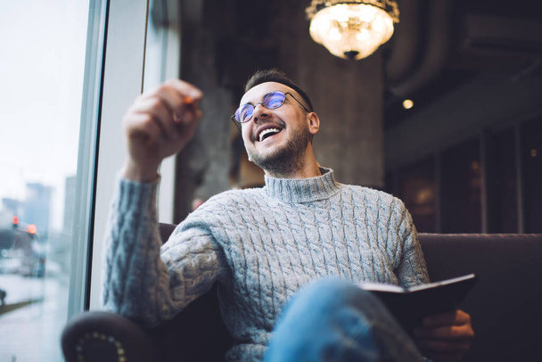 Cheerful bearded male in casual wear and glasses smiling and looking away while sitting in comfortable armchair and writing information - Zdjęcie, obraz