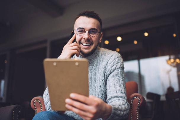 Ein bärtiger Mann in lässiger Kleidung und Brille, der bei einem Telefonat im Café lächelnd in die Kamera blickt - Foto, Bild