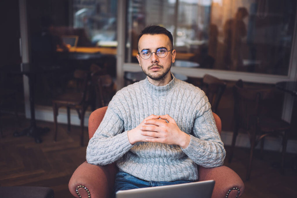 High angle of confident bearded male in casual outfit and eyeglasses sitting in armchair and looking at camera while browsing laptop - Фото, зображення