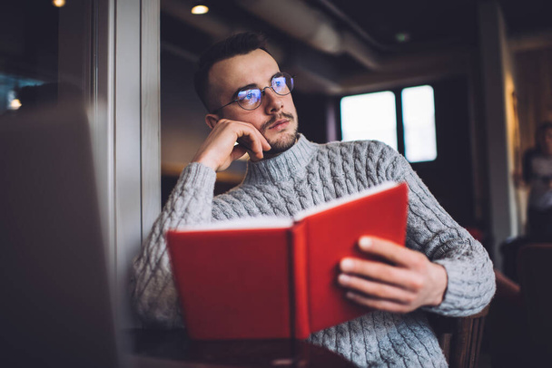 Serious young bearded male freelancer in casual clothes and eyeglasses sitting in modern cafeteria and looking away while resting with book - Fotografie, Obrázek
