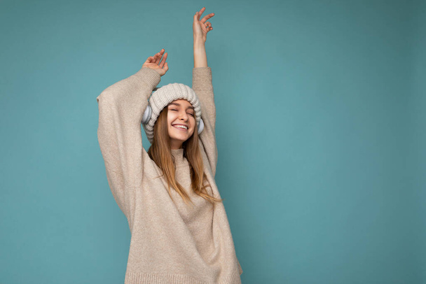 Photo of beautiful happy smiling young blonde woman wearing beige winter sweater and hat isolated over blue background wearing white wireless bluetooth headphones listening to cool music dancing and - Fotografie, Obrázek