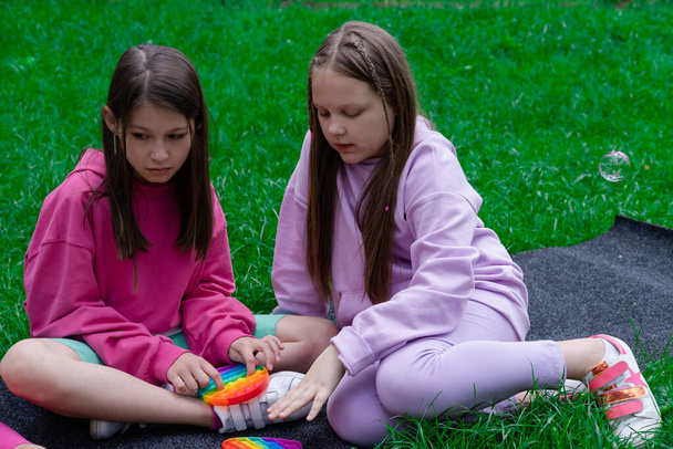 İki genç kız, modaya uygun duyusal oyuncaklarla oynuyorlar çimlerin üzerinde oynuyorlar. Popo kabarcığı esnek, hareketli duyusal oyuncağı itin. Stres ve endişe giderici. Stresli çocuklar ve yetişkinler için moda silikon oyunu. - Fotoğraf, Görsel