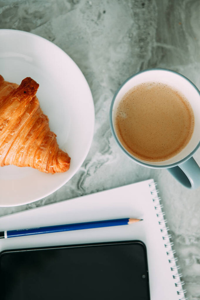 business breakfast in a light key, coffee and a croissant, a white notebook, a black phone and a blue pencil - Foto, Bild