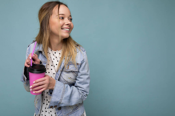 Attractive young happy smiling blonde woman wearing everyday stylish clothes isolated over colourful background wall holding paper cup for cutout drinking tea looking to the side - 写真・画像