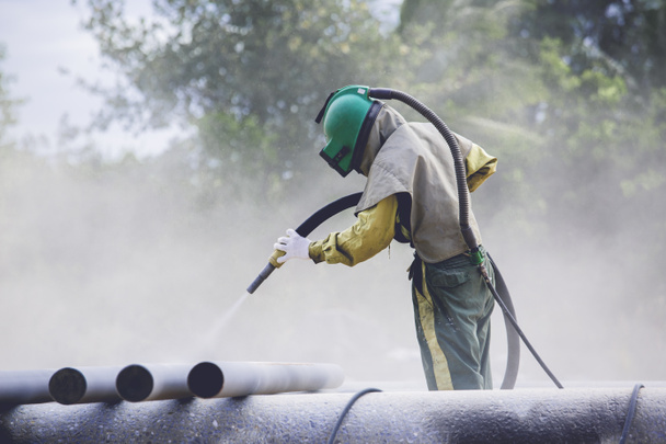 Mannelijke werknemer zandstralen stofproces reiniging pijpleiding oppervlak op staal voor het schilderen in de fabriek. - Foto, afbeelding