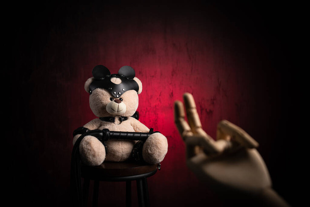 un osito de peluche de juguete con un látigo, vestido con cinturones de cuero y una máscara, un accesorio para juegos BDSM con una mano de madera y un gesto sexual - Foto, Imagen