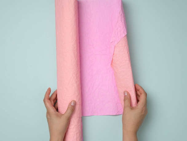 rouleau rose de papier d'emballage dans les mains féminines sur un fond bleu, vue de dessus - Photo, image