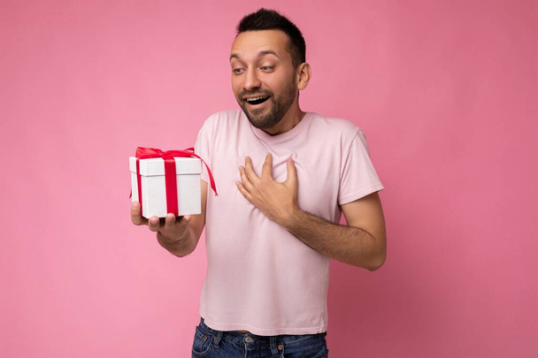 Shot of happy shocked brunet νεαρός άνδρας με γενειάδα απομονωμένος πάνω από ροζ φόντο τοίχο φορώντας ροζ t-shirt κρατώντας λευκό κουτί δώρου με κόκκινη κορδέλα και κοιτάζοντας προς το παρόν - Φωτογραφία, εικόνα
