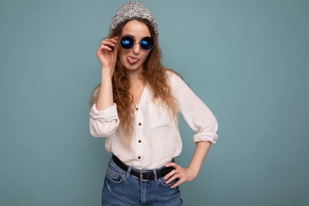 Foto shot van aantrekkelijke jonge krullende vrouw geïsoleerd over blauwe achtergrond dragen casual witte blouse en stijlvolle bril kijken naar de camera en plezier hebben - Foto, afbeelding