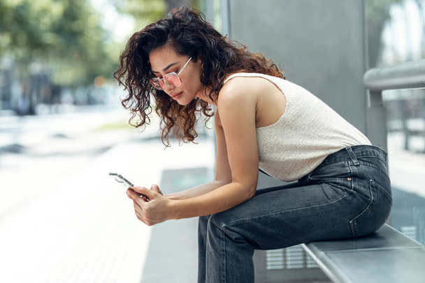 Aufnahme einer lächelnden jungen Geschäftsfrau mit ihrem Handy, die auf einer Bank auf der Straße sitzt. - Foto, Bild