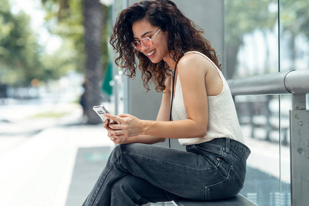 Prise de vue de jeune femme d'affaires souriante utilisant son téléphone portable assis sur un banc dans la rue. - Photo, image