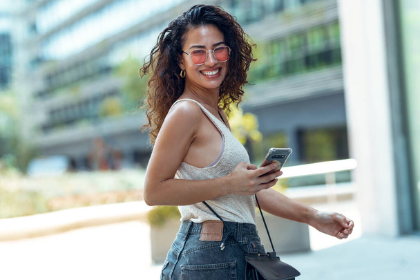 Shot of beautiful young business woman using her mobile phone while looking at camera walking down the street. - Foto, Imagen