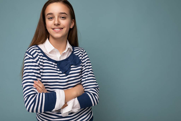 Photo shot of beautiful positive smiling brunette little girl wearing striped longsleeve standing isolated over blue background wall crossing hands and looking at camera - Photo, Image
