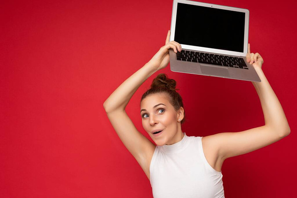 schöne erstaunt faszinierende glücklich blonde Frau mit gerafften Haaren, die in die Kamera schaut, Computer-Laptop mit leerem Monitor mit Attrappe und Kopierraum trägt weißes T-Shirt - Foto, Bild