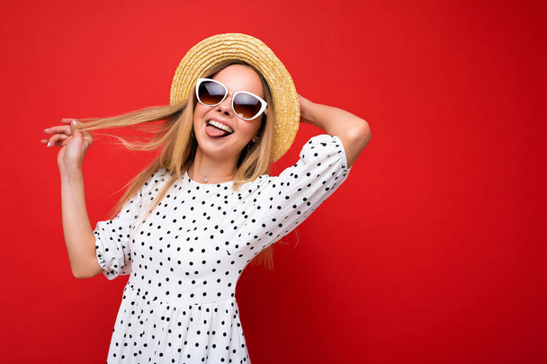 Atraente jovem loira feliz vestindo roupas elegantes todos os dias e óculos de sol modernos isolados na parede de fundo colorido olhando para a câmera se divertindo e mostrando língua - Foto, Imagem