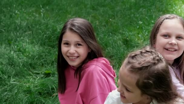 Kolme kaunista onnellista teini-ikäistä tyttöä värikkäissä vaatteissa istumassa vihreällä ruoholla puistossa. Kesällä. Onnellisia lapsia. Lomat, kesäloma. Siskot, parhaat ystävät, bff, sisaruus. hidastettuna. - Materiaali, video