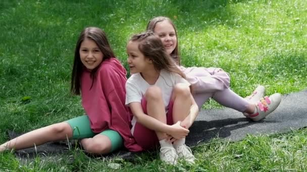 Parktaki yeşil çimlerde oturan renkli elbiseli üç güzel ergen kız. Yaz zamanı. Mutlu çocuklar. Tatil, yaz tatili. Kız kardeşler, en iyi arkadaşlar, en iyi arkadaşlar, kardeşlik. yavaş çekim. - Video, Çekim