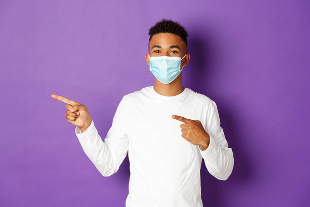 Konzept von Coronavirus, Quarantäne und Lebensstil. Fröhlicher afrikanisch-amerikanischer Mann in medizinischer Maske zeigt Werbung, zeigt nach links und lächelt, steht über lila Hintergrund - Foto, Bild