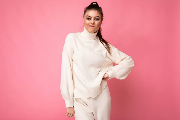 Young beautiful self-confident sexy stylish brunette female person wearing casual white sweater poising isolated on pink background wall - 写真・画像