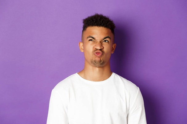 Close-up van de lastige jonge Afrikaans-Amerikaanse man, omhoog kijkend en grimmig verbijsterd, moeilijke keuze hebbend, staande over paarse achtergrond - Foto, afbeelding