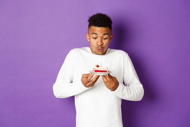 Happy african-american man celebrating birthday, blowing candle on a cake and making wish, standing over purple background - Photo, image