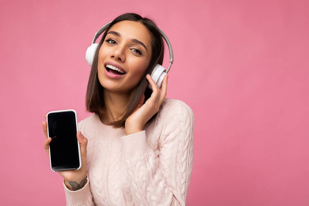 セクシーな美しいかわいい若いブルネットの女性の肖像ピンクのセーターを身に着けているピンクの背景の壁に白いブルートゥースワイヤレスヘッドフォンを身に着けていると良い音楽を聞いて、モバイルを示す - 写真・画像