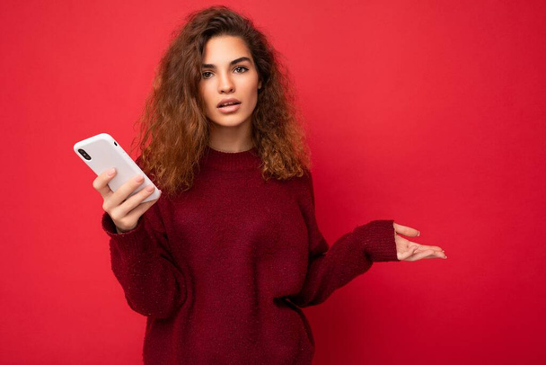 Фото довольно спрашивает расстроенной молодой женщины с кудрявыми волосами носить темно-красный свитер изолированы на красном фоне держа мобильный телефон глядя на камеру и не понимая - Фото, изображение