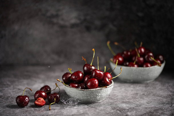 Verter agua sobre cerezas rojas amargas y dulces frescas en dos cuencos de escándalo grises sobre fondo gris y hacer gotas de agua - Foto, imagen