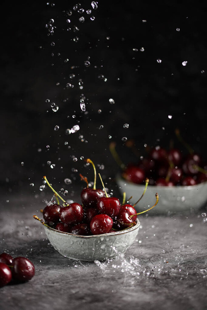 Verter agua sobre cerezas rojas amargas y dulces frescas en dos cuencos de escándalo grises sobre fondo gris oscuro y hacer gotas de agua - Foto, imagen