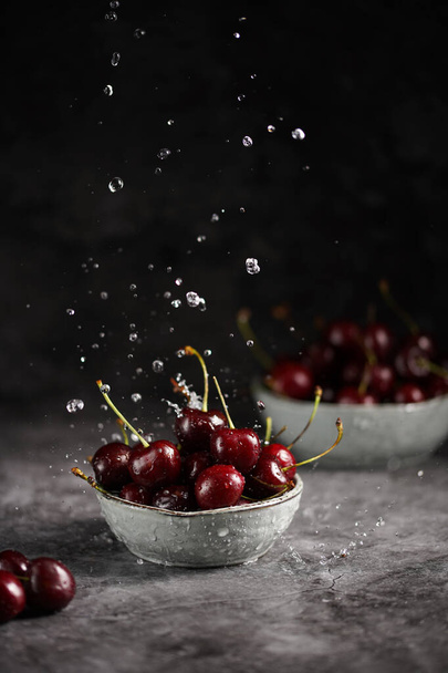 Pouring water over fresh sweet and sour dark red cherries in two grey scandi bowls on dark grey background and making water drops - Fotó, kép