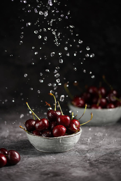 Versare acqua su ciliegie fresche dolci e acide rosso scuro in due ciotole grigie scandi su fondo grigio scuro e fare gocce d'acqua - Foto, immagini