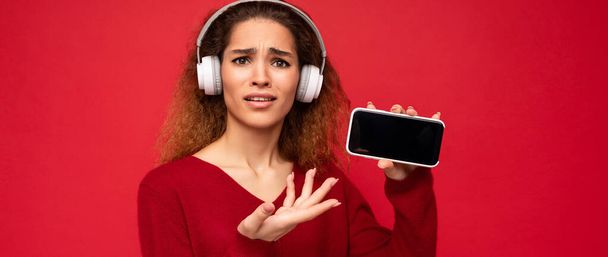 Attraktive unzufriedene schockierte junge brünette lockige Frau trägt dunkelroten Pullover isoliert auf rotem Hintergrund mit weißen Headsets Musik hören und zeigt moderne Handy mit leerem Bildschirm für - Foto, Bild