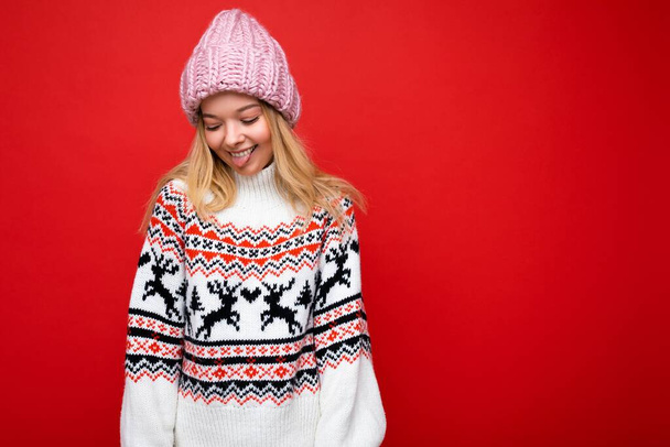 写真のかなり肯定的な笑顔若いですブロンド女性絶縁以上赤背景壁身に着けています冬のセーターとピンクの帽子見下す - 写真・画像