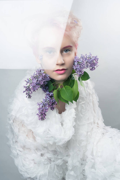 Mujer rubia joven con lilas en un vestido blanco. Retrato de estudio sobre fondo blanco a través del vidrio - Foto, imagen