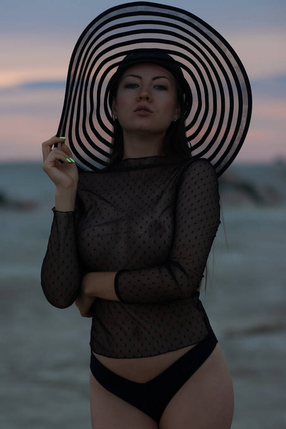 Confident attractive fashionable young female in provocative translucent swimwear and hat looking at camera while resting on beach at sunset time - Photo, Image