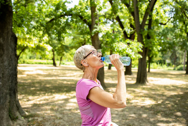 Starsza kaukaska kobieta robi sobie przerwę i pije wodę po długim treningu biegowym w przyrodzie. - Zdjęcie, obraz