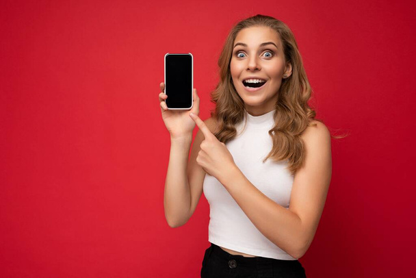 Потрясенная красивая молодая блондинка с открытым ртом в белой футболке, изолированной на красном фоне с копировальным пространством, держа смартфон, показывающий телефон с пустым экраном для макета, глядя на - Фото, изображение