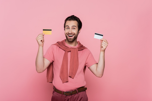 Positive man holding credit cards and looking at camera isolated on pink  - Photo, Image