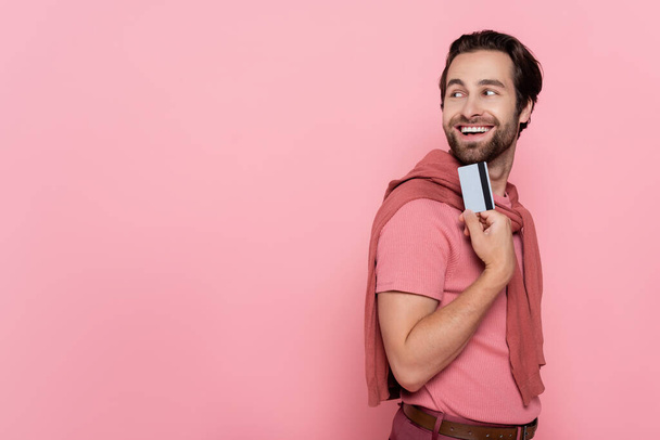 Brunette man smiling while holding credit card and looking away isolated on pink  - Foto, Bild