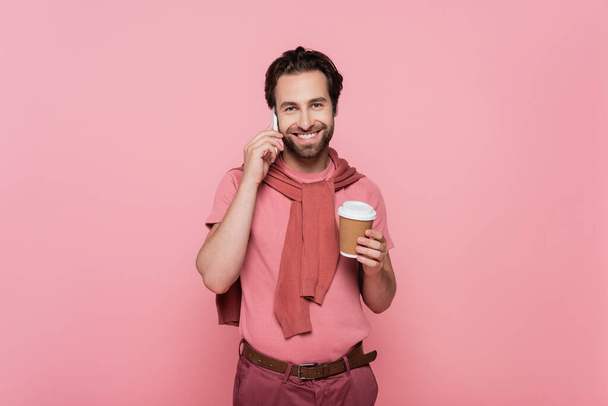 Positive man looking at camera while talking on mobile phone and holding coffee to go isolated on pink  - Foto, afbeelding