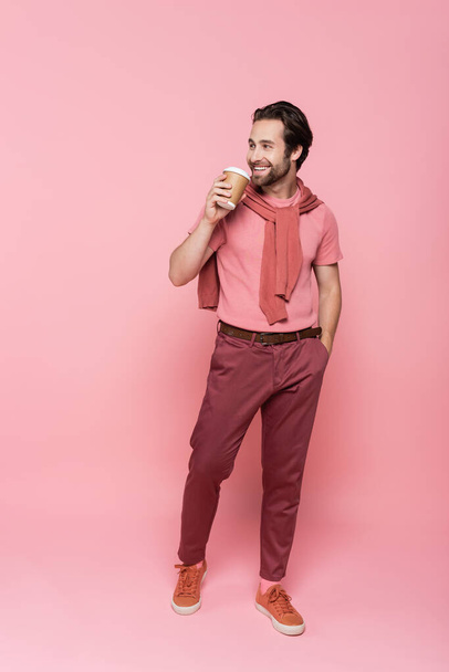 Smiling man with takeaway drink holding hand in pocket on pink background - Фото, изображение