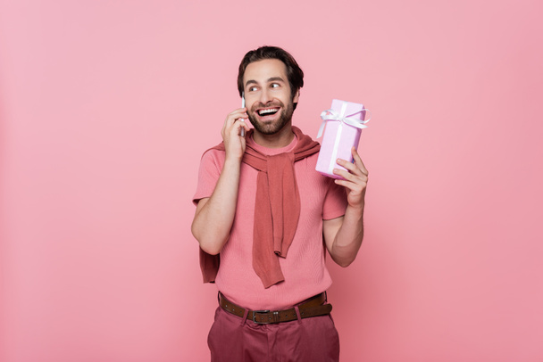 Excited man talking on mobile phone and holding present isolated on pink  - Zdjęcie, obraz