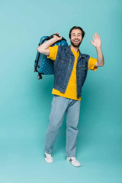 Happy traveler in denim vest met koffer en zwaaiende hand op blauwe achtergrond  - Foto, afbeelding