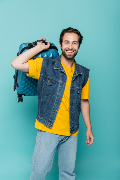 Cheerful tourist holding baggage isolated on blue  - Photo, Image