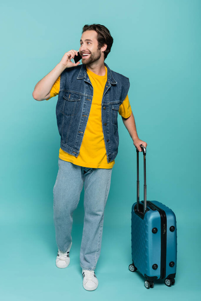Smiling tourist talking on mobile phone and holding suitcase on blue background  - Fotografie, Obrázek